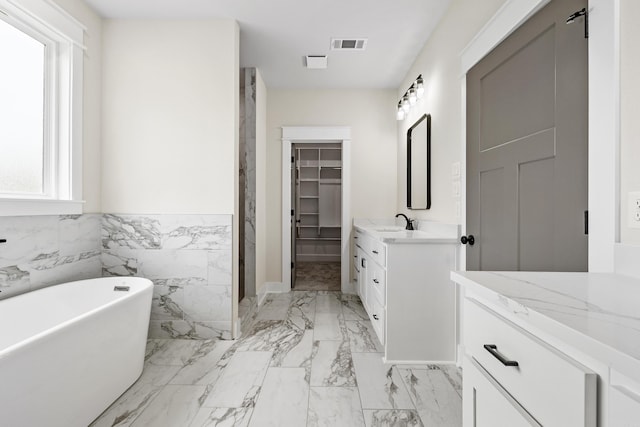 full bath featuring vanity, a freestanding tub, a spacious closet, marble finish flooring, and wainscoting