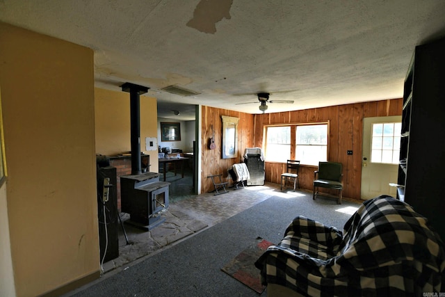 carpeted living room featuring a textured ceiling, visible vents, wood walls, ceiling fan, and a wood stove