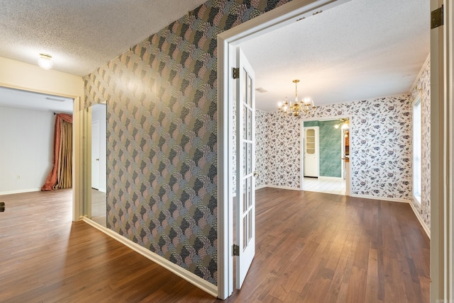 hall featuring a textured ceiling, wallpapered walls, an inviting chandelier, and wood-type flooring