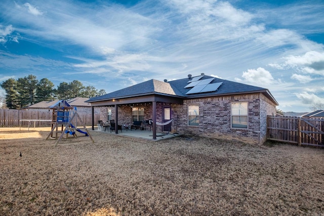 back of property featuring a playground, a fenced backyard, solar panels, brick siding, and a patio area