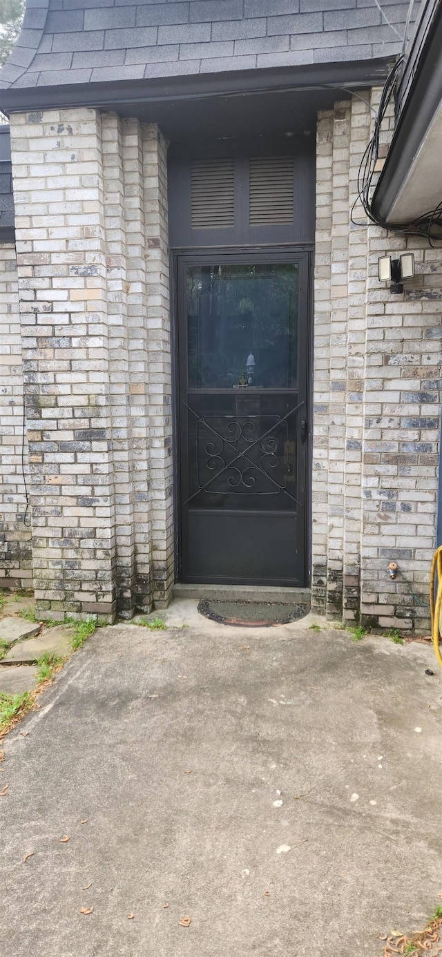 view of exterior entry with a shingled roof and brick siding