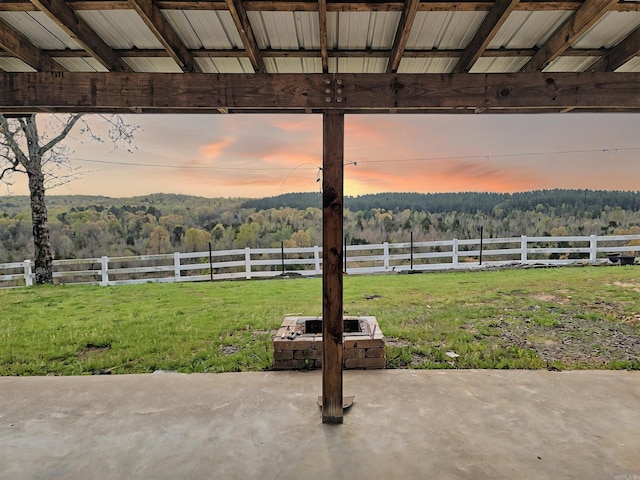 patio terrace at dusk with a lawn, a wooded view, fence, and an outdoor fire pit