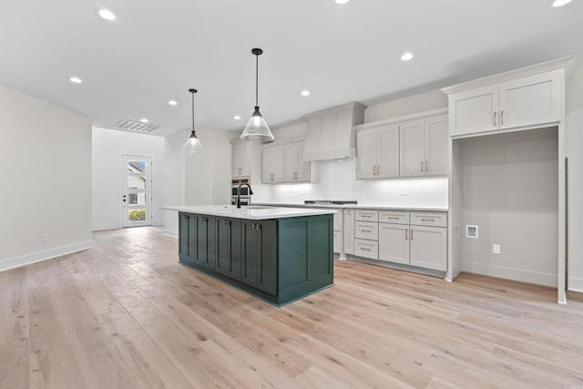 kitchen featuring light countertops, a kitchen island with sink, cooktop, light wood-style floors, and custom range hood