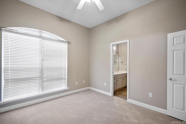 unfurnished bedroom with ensuite bathroom, light colored carpet, a ceiling fan, multiple windows, and baseboards