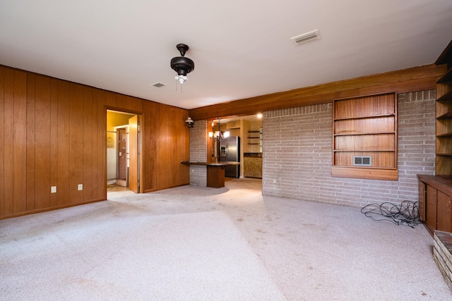 unfurnished living room featuring light colored carpet, wood walls, a notable chandelier, and visible vents