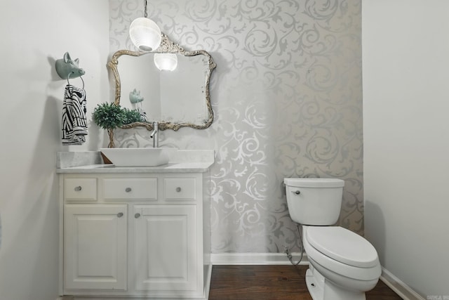half bath featuring vanity, baseboards, wallpapered walls, toilet, and wood finished floors
