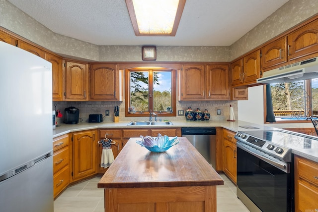 kitchen featuring a wealth of natural light, brown cabinetry, under cabinet range hood, a sink, and stainless steel appliances
