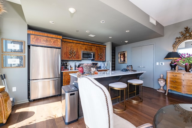 kitchen featuring tasteful backsplash, a center island with sink, brown cabinets, stainless steel appliances, and wood finished floors