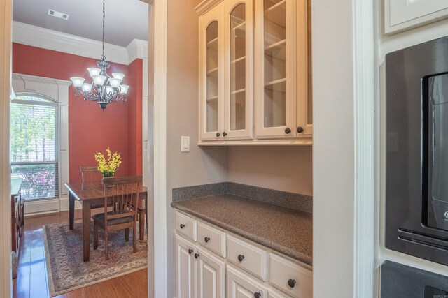 kitchen with an inviting chandelier, glass insert cabinets, crown molding, dark countertops, and wood finished floors