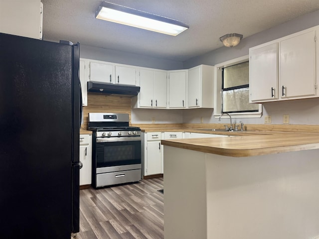 kitchen with wooden counters, a peninsula, under cabinet range hood, stainless steel gas range oven, and freestanding refrigerator