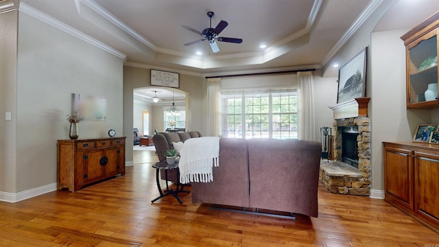 living area featuring a raised ceiling, a stone fireplace, a ceiling fan, ornamental molding, and light wood-type flooring