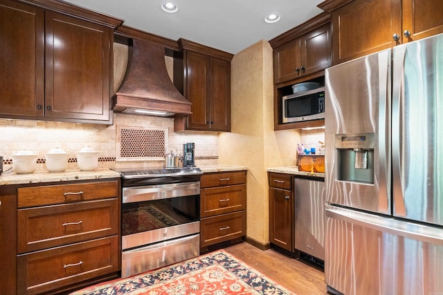 kitchen with backsplash, light wood-style floors, light stone countertops, appliances with stainless steel finishes, and custom range hood