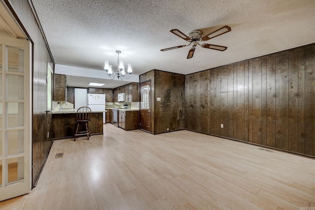 unfurnished living room featuring wood walls, light wood-style flooring, and ceiling fan with notable chandelier