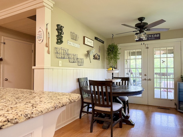 dining area featuring light wood finished floors, wainscoting, a ceiling fan, and french doors