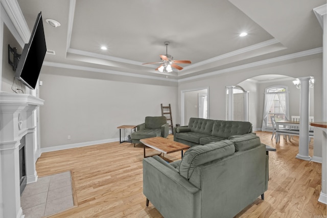 living room featuring a fireplace with flush hearth, ornate columns, a tray ceiling, arched walkways, and ceiling fan