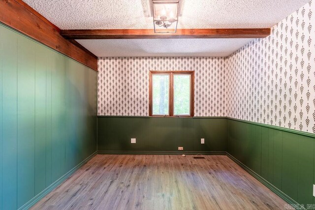 empty room featuring wood finished floors, wallpapered walls, a textured ceiling, a wainscoted wall, and beam ceiling