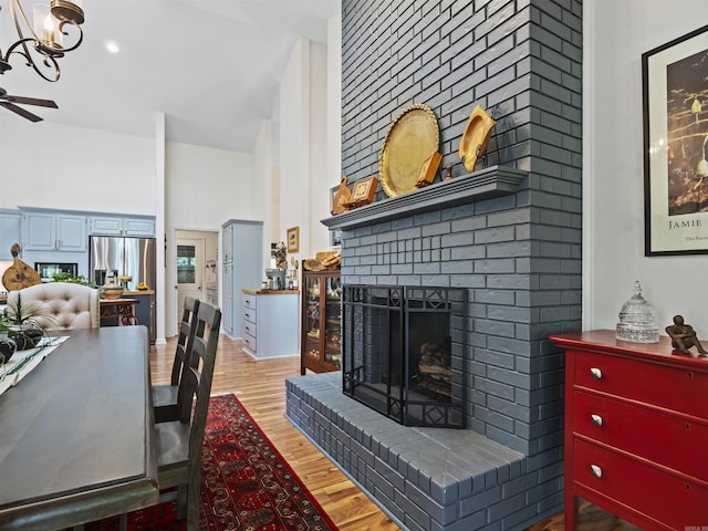 dining room with light wood-type flooring, a high ceiling, a fireplace, and ceiling fan with notable chandelier