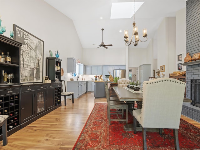 dining space featuring light wood finished floors, a skylight, a fireplace, high vaulted ceiling, and ceiling fan with notable chandelier