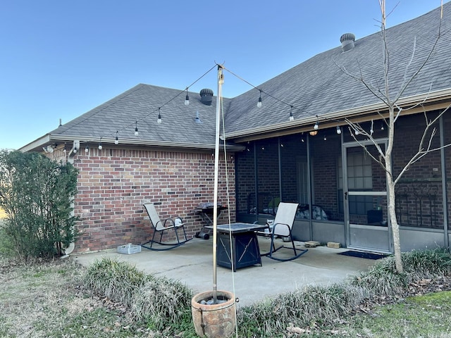 back of house featuring a sunroom, an outdoor fire pit, brick siding, a patio area, and roof with shingles