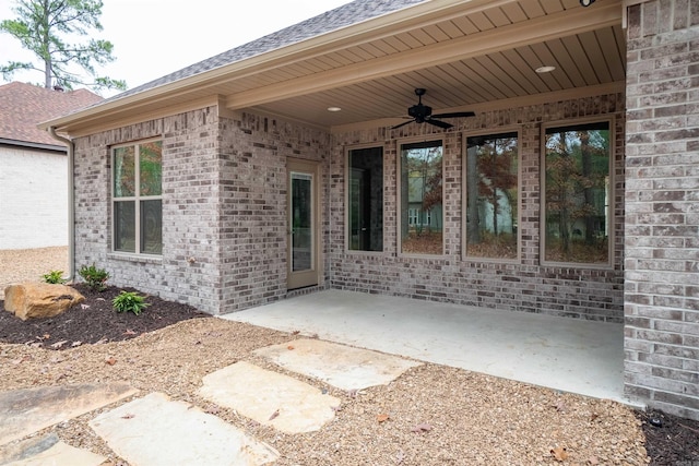 view of exterior entry with a ceiling fan, roof with shingles, a patio area, and brick siding
