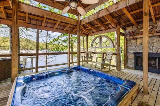 wooden deck featuring hot tub deck surround, a ceiling fan, an outdoor stone fireplace, and a water view