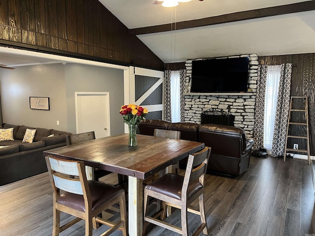 dining room featuring wood walls, wood finished floors, a stone fireplace, vaulted ceiling with beams, and a ceiling fan