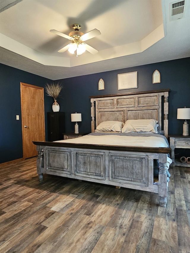 bedroom with a ceiling fan, a tray ceiling, visible vents, and dark wood-type flooring