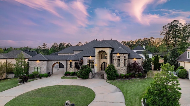 french country inspired facade featuring curved driveway, a shingled roof, a yard, and brick siding