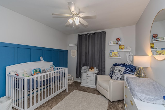 bedroom with ceiling fan, wainscoting, a crib, carpet, and a decorative wall