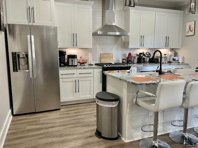 kitchen with appliances with stainless steel finishes, white cabinetry, light stone counters, light wood finished floors, and wall chimney exhaust hood