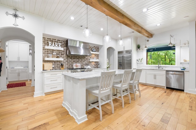 kitchen with wall chimney range hood, appliances with stainless steel finishes, arched walkways, open shelves, and light wood-style floors