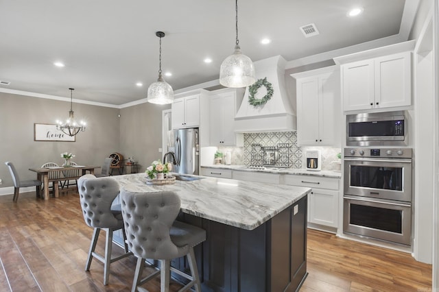 kitchen with a kitchen island with sink, premium range hood, stainless steel appliances, white cabinetry, and backsplash
