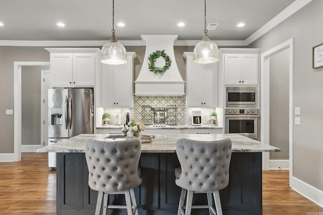 kitchen featuring appliances with stainless steel finishes, premium range hood, white cabinetry, tasteful backsplash, and a center island with sink
