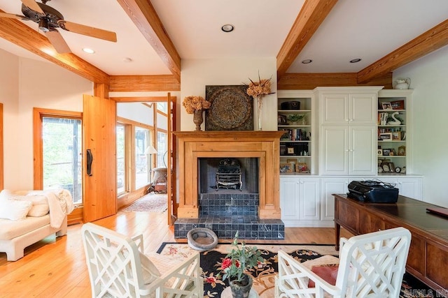 living area featuring a ceiling fan, beam ceiling, light wood-style flooring, and a tile fireplace