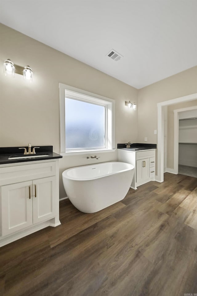 full bath featuring visible vents, wood finished floors, a freestanding bath, two vanities, and a sink