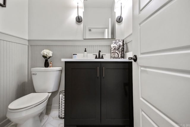 half bathroom featuring wainscoting, vanity, toilet, marble finish floor, and a decorative wall
