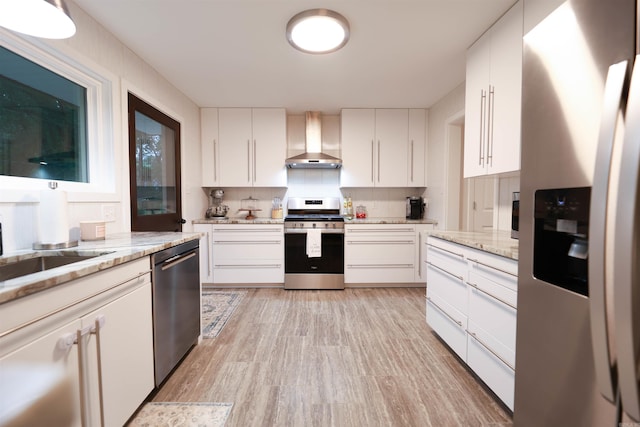 kitchen with light stone countertops, white cabinetry, light wood finished floors, stainless steel appliances, and wall chimney exhaust hood