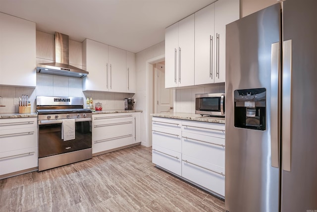 kitchen with wall chimney range hood, decorative backsplash, light stone countertops, white cabinets, and stainless steel appliances