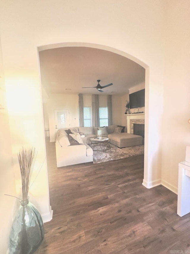 living room featuring a ceiling fan, wood finished floors, a stone fireplace, baseboards, and arched walkways
