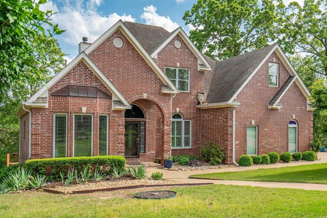 view of front facade featuring brick siding, roof with shingles, a front yard, and a chimney