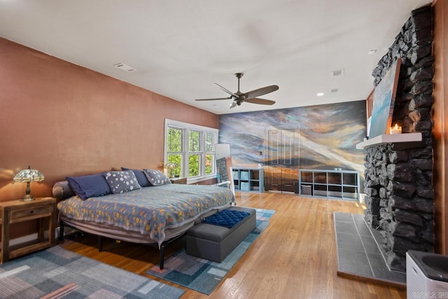 bedroom with ceiling fan, hardwood / wood-style flooring, a stone fireplace, and visible vents