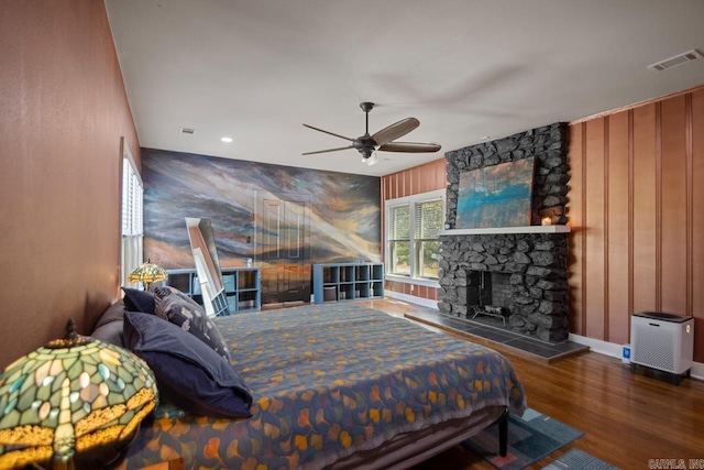 bedroom with visible vents, an accent wall, wood finished floors, baseboards, and a stone fireplace
