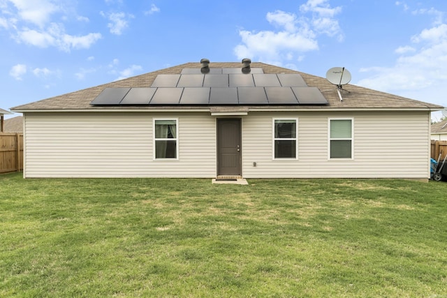 rear view of house with roof mounted solar panels, fence, a shingled roof, and a yard