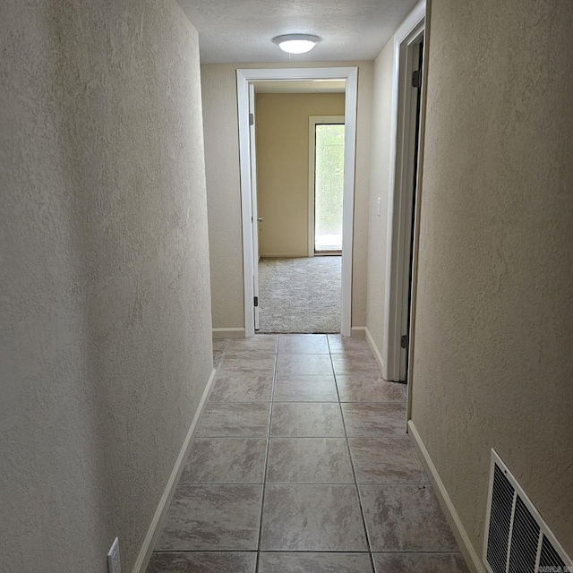 hall with light tile patterned flooring, a textured wall, visible vents, and baseboards