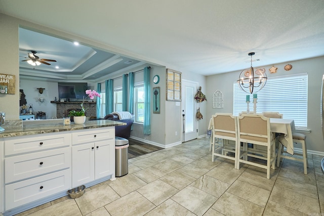 kitchen featuring ceiling fan with notable chandelier, light stone counters, a brick fireplace, white cabinetry, and baseboards
