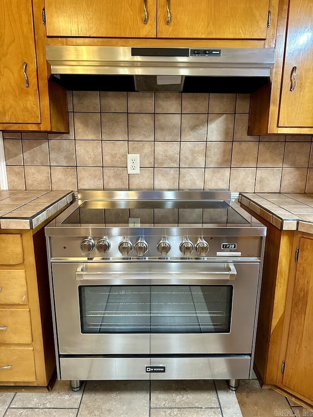 details featuring high end stainless steel range, tile counters, brown cabinets, and backsplash