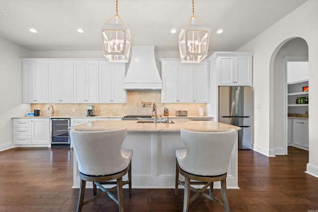 kitchen with beverage cooler, freestanding refrigerator, a kitchen island with sink, decorative backsplash, and custom exhaust hood