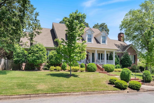 new england style home with a chimney, brick siding, covered porch, a shingled roof, and a front lawn
