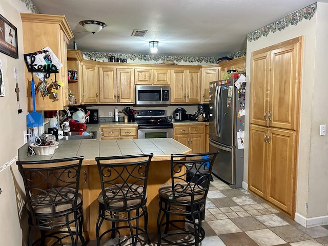 kitchen with appliances with stainless steel finishes, a peninsula, tile countertops, a sink, and a kitchen breakfast bar