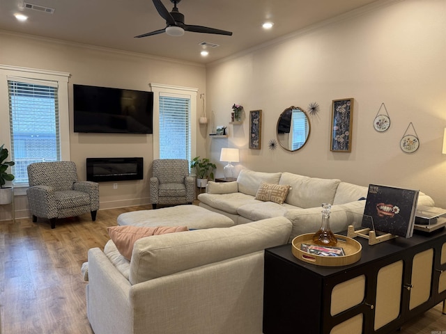 living area with ceiling fan, wood finished floors, crown molding, a glass covered fireplace, and baseboards